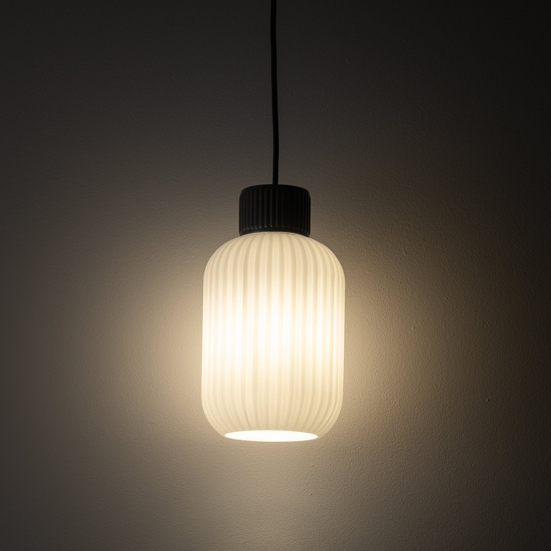 Na obrazie widoczna jest wisząca lampa z żarówką w środku, emitująca ciepłe światło. Klosz lampy ma pionowe żłobienia i jest zawieszony na czarnym przewodzie.