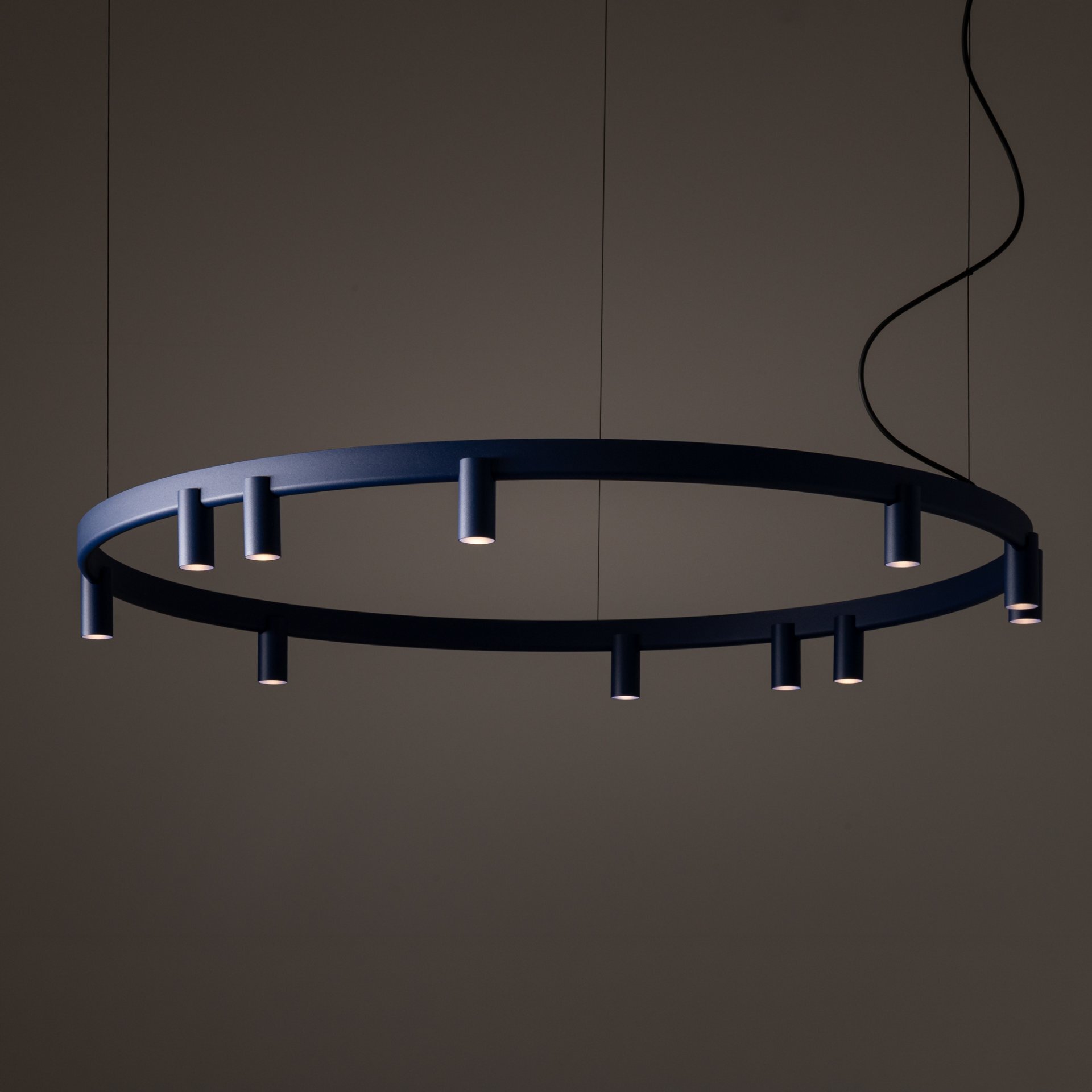 Na obrazie widoczna jest nowoczesna lampa wisząca w kształcie okręgu z kilkoma źródłami światła skierowanymi w dół. Lampa jest zawieszona na cienkich przewodach, a jej kolor to ciemny odcień, prawdopodobnie czarny lub granatowy.