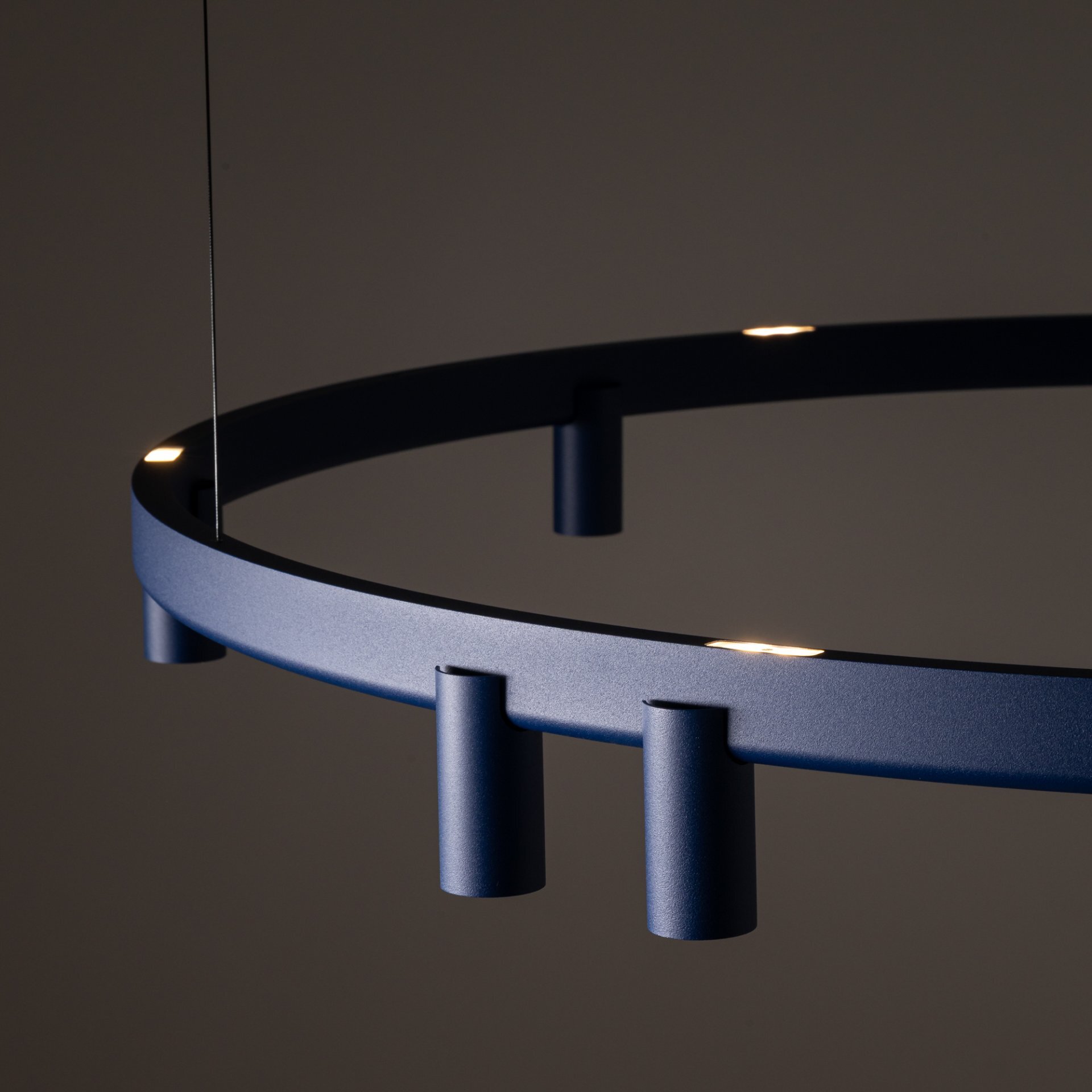Na obrazie widoczna jest nowoczesna lampa wisząca o okrągłym kształcie, wykonana z metalu w ciemnym kolorze. Lampa posiada kilka punktów świetlnych rozmieszczonych równomiernie na obręczy.