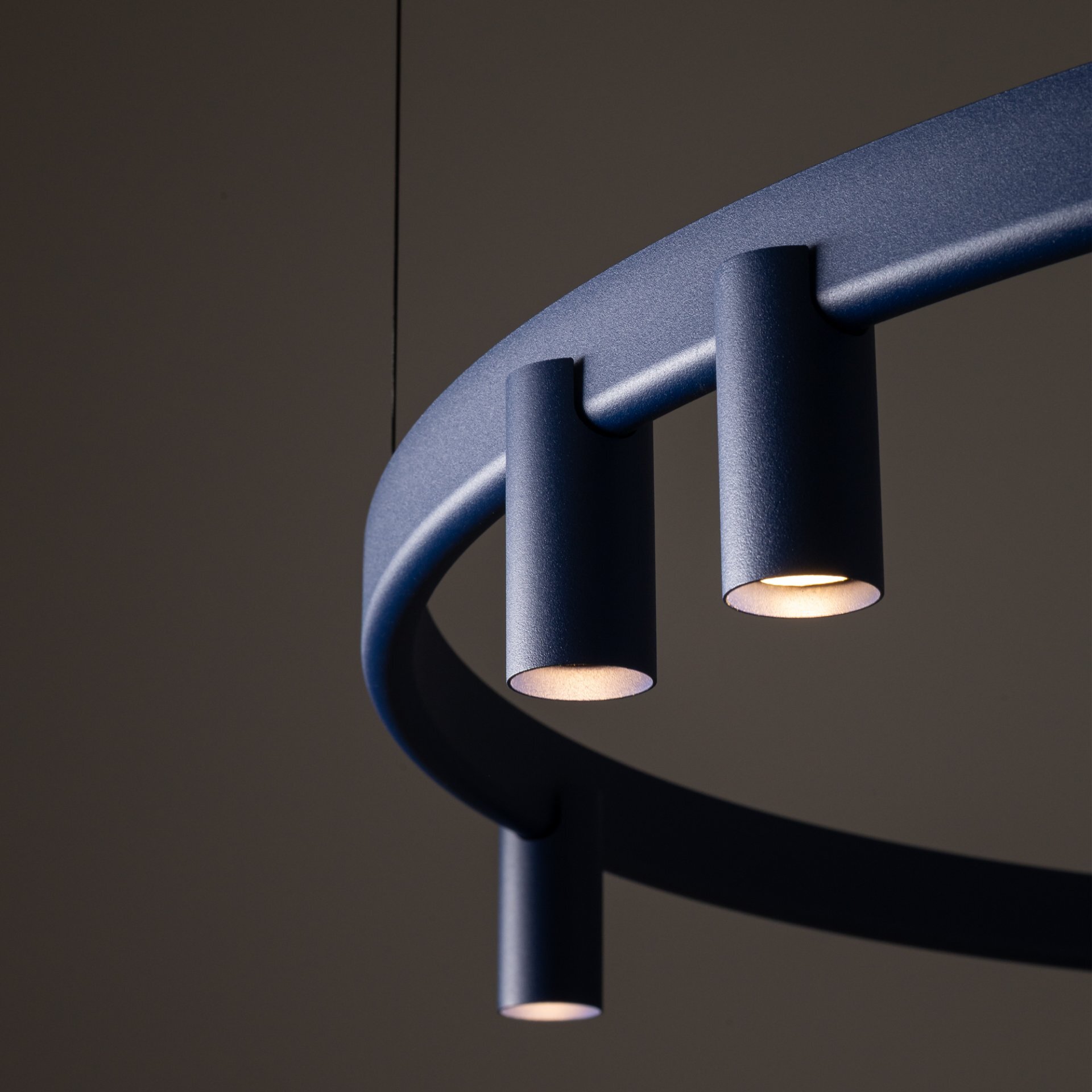 Na obrazie widoczna jest nowoczesna lampa wisząca o okrągłym kształcie z trzema cylindrycznymi źródłami światła. Lampa ma ciemny kolor i jest zawieszona na cienkim przewodzie.