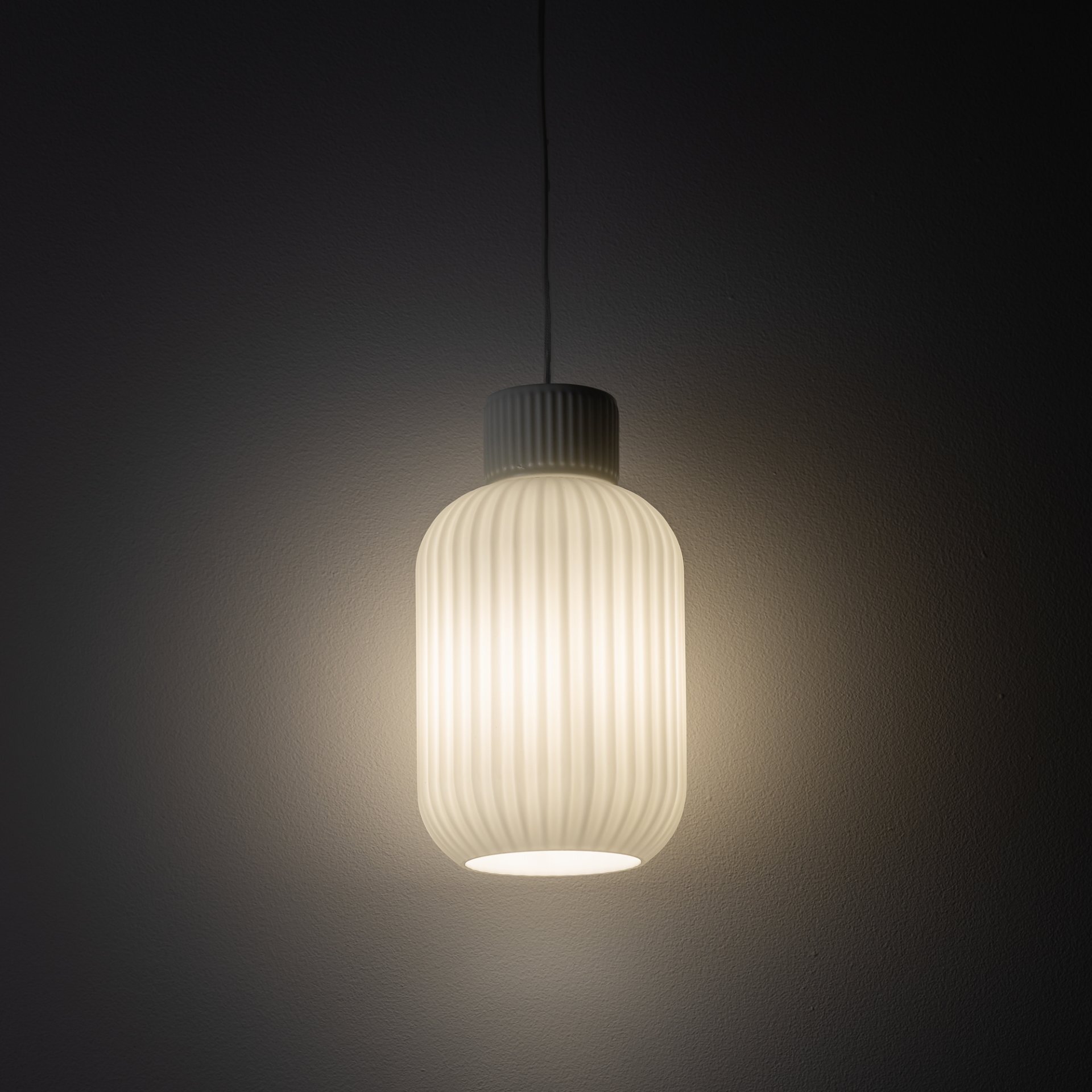 Na obrazie widoczna jest wisząca lampa o cylindrycznym kształcie z pionowymi żłobieniami, emitująca ciepłe, rozproszone światło. Tło jest ciemne, co podkreśla jasność lampy.