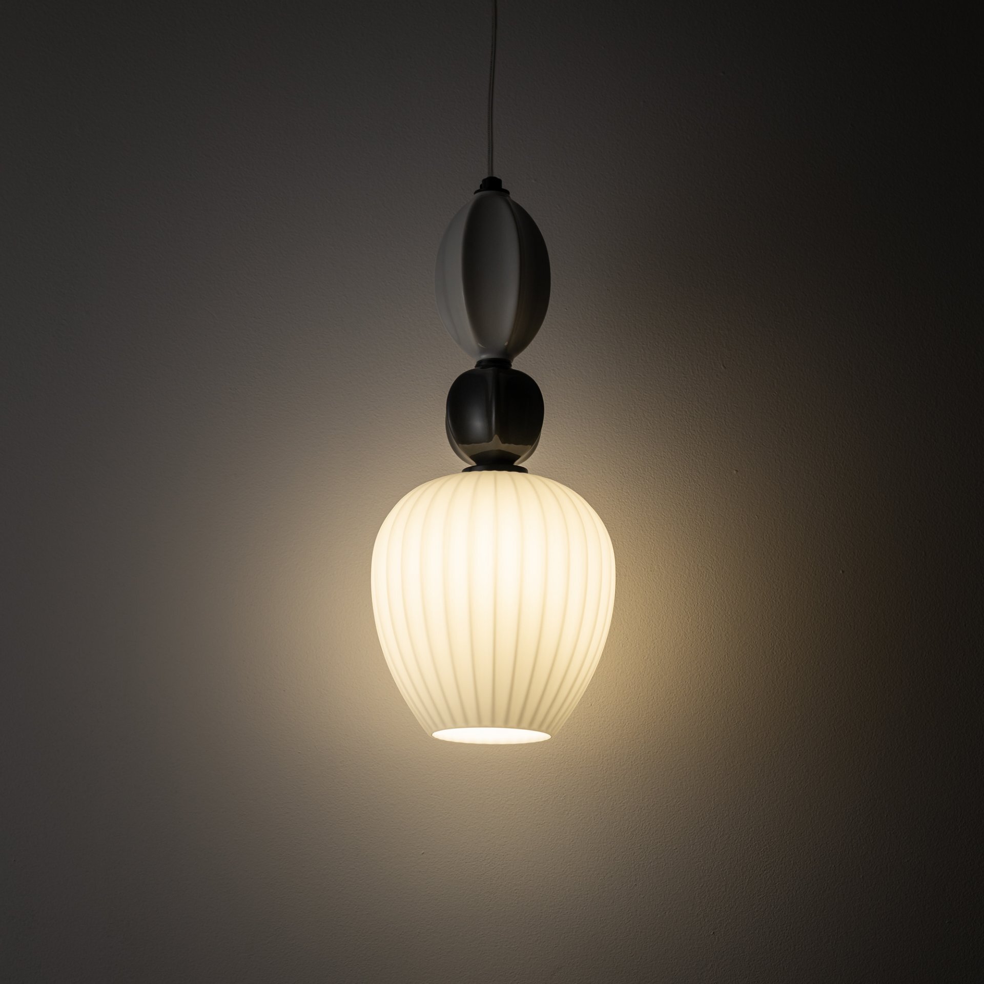 Na obrazie widoczna jest wisząca lampa o białym, żebrowanym kloszu, emitująca ciepłe światło. Lampa zawieszona jest na ciemnym kablu, a jej górna część składa się z dwóch ozdobnych elementów.