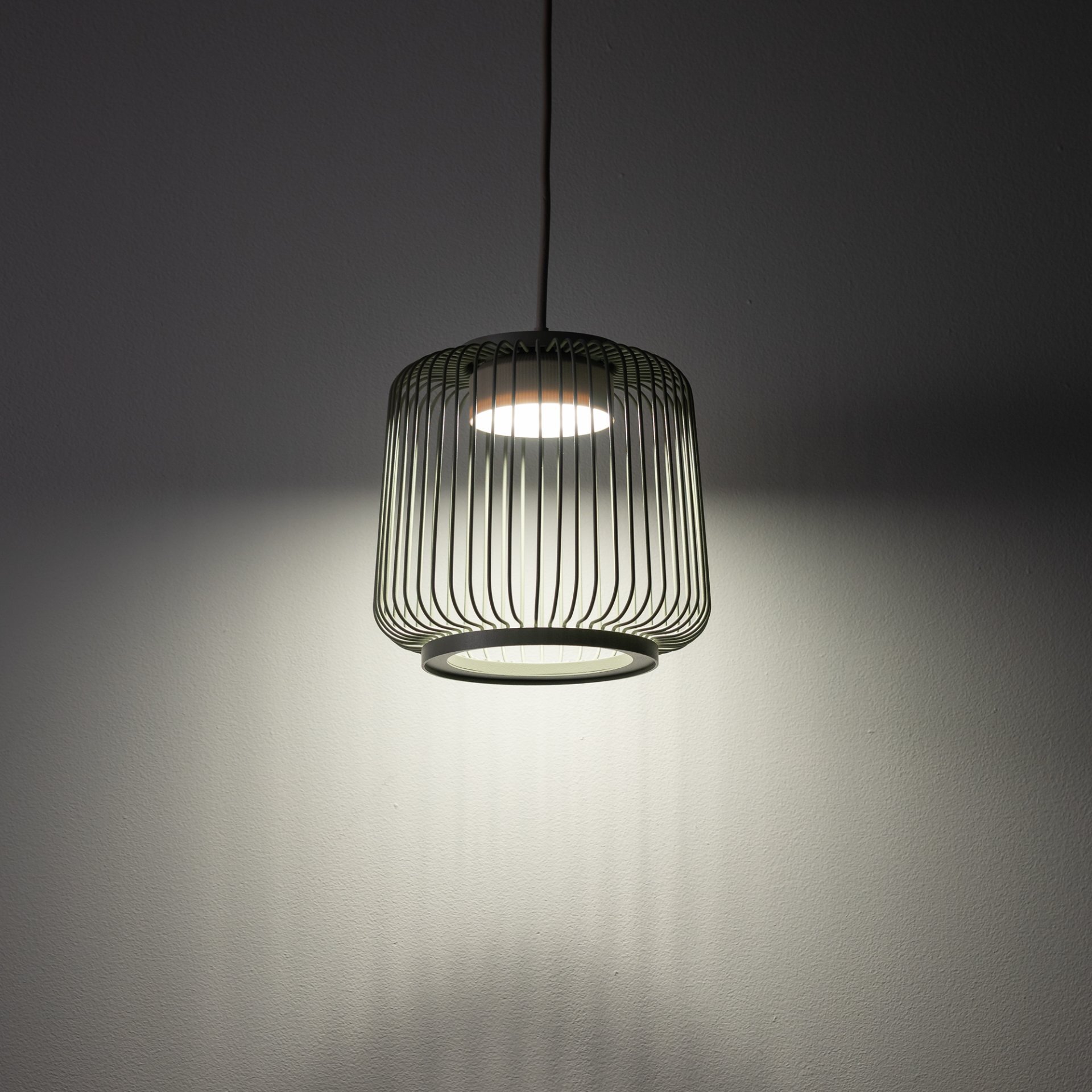 Lampa wisząca z metalowym kloszem w formie ażurowej konstrukcji, emitująca światło na ścianę. Światło tworzy delikatny wzór cieni na powierzchni.