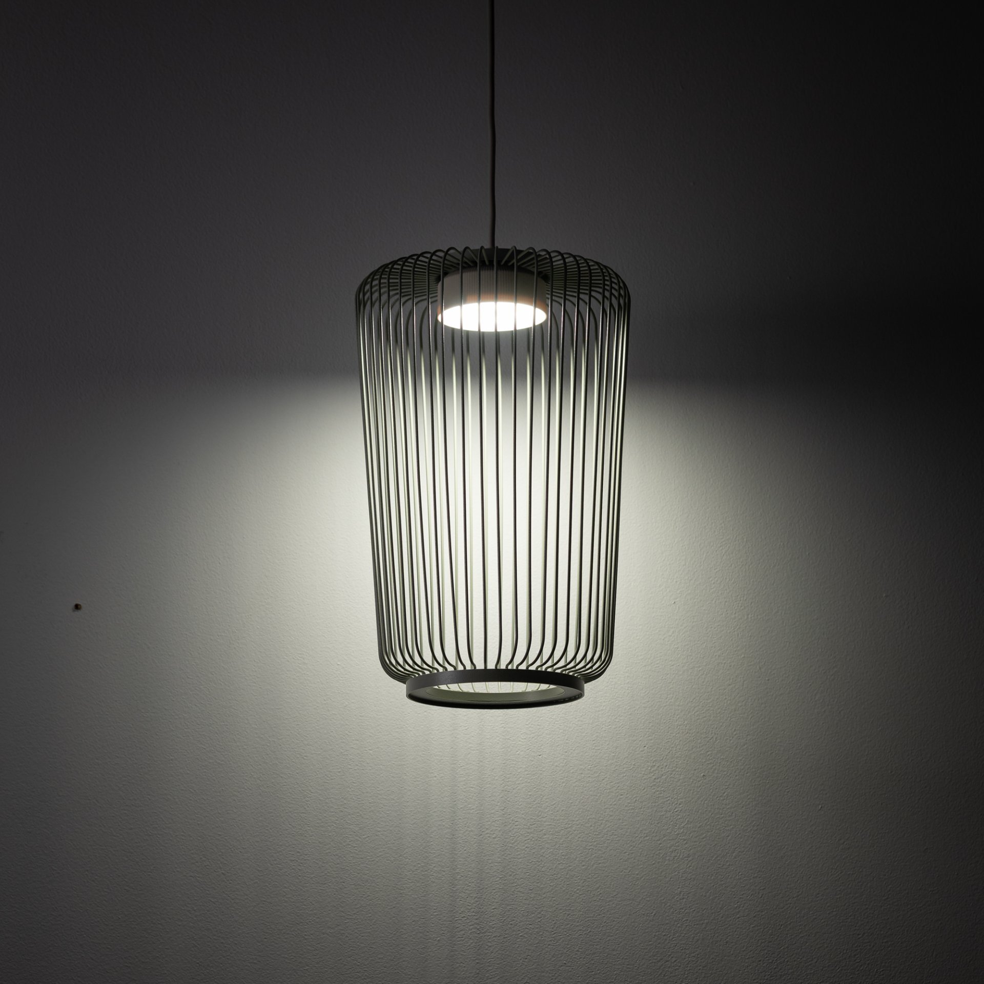 Na obrazie widoczna jest wisząca lampa z metalowym kloszem o cylindrycznym kształcie, emitująca światło na ścianę. Lampa jest włączona, a jej światło tworzy delikatny efekt na tle ciemnej ściany.