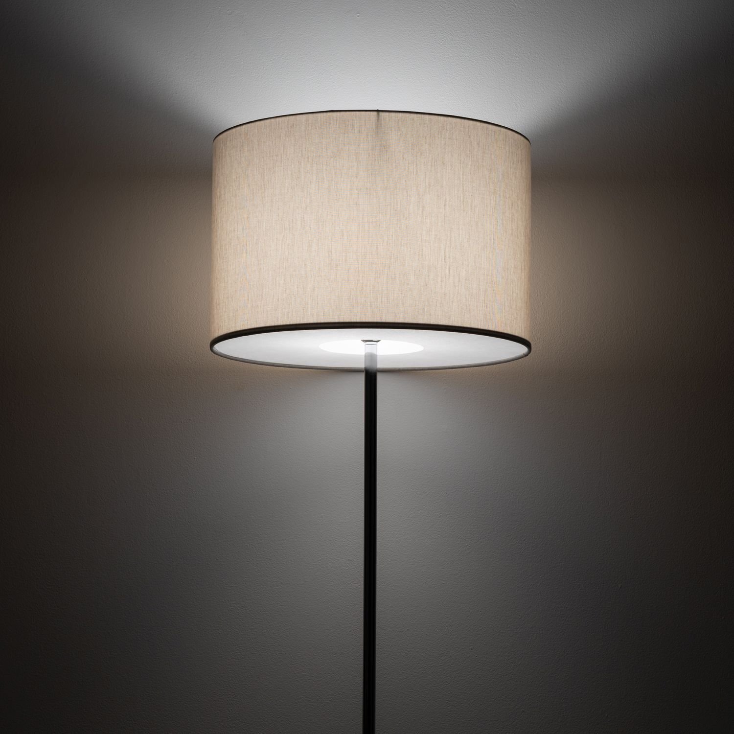 Lampa podłogowa z jasnym, cylindrycznym abażurem, emitująca rozproszone światło na ścianę. Czarny, smukły trzon lampy jest widoczny poniżej abażuru.