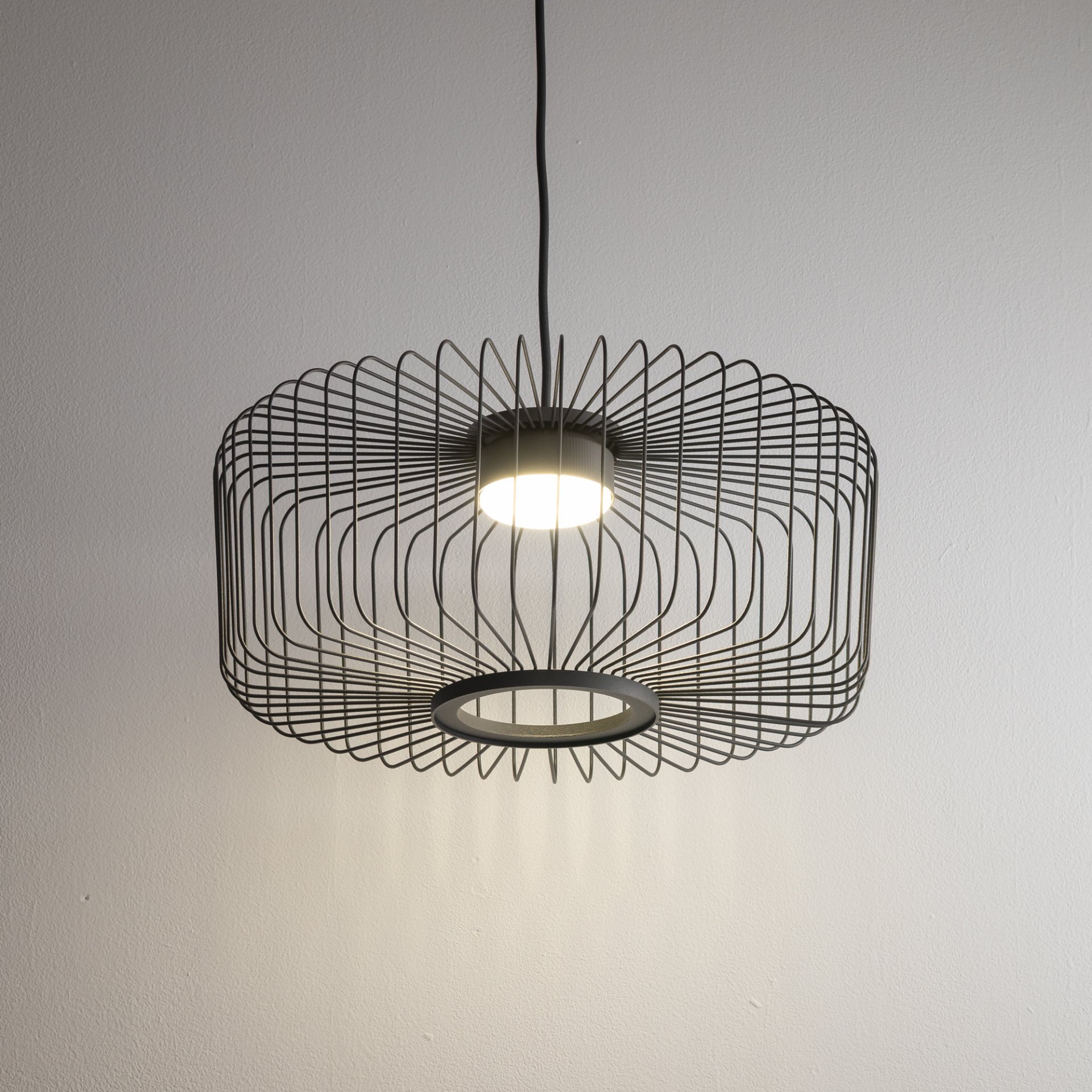 Lampa wisząca z metalowym kloszem o geometrycznym wzorze, emitująca światło z centralnego punktu. Klosz ma otwartą konstrukcję z cienkich prętów, co tworzy efektowny wzór cieni na ścianie.