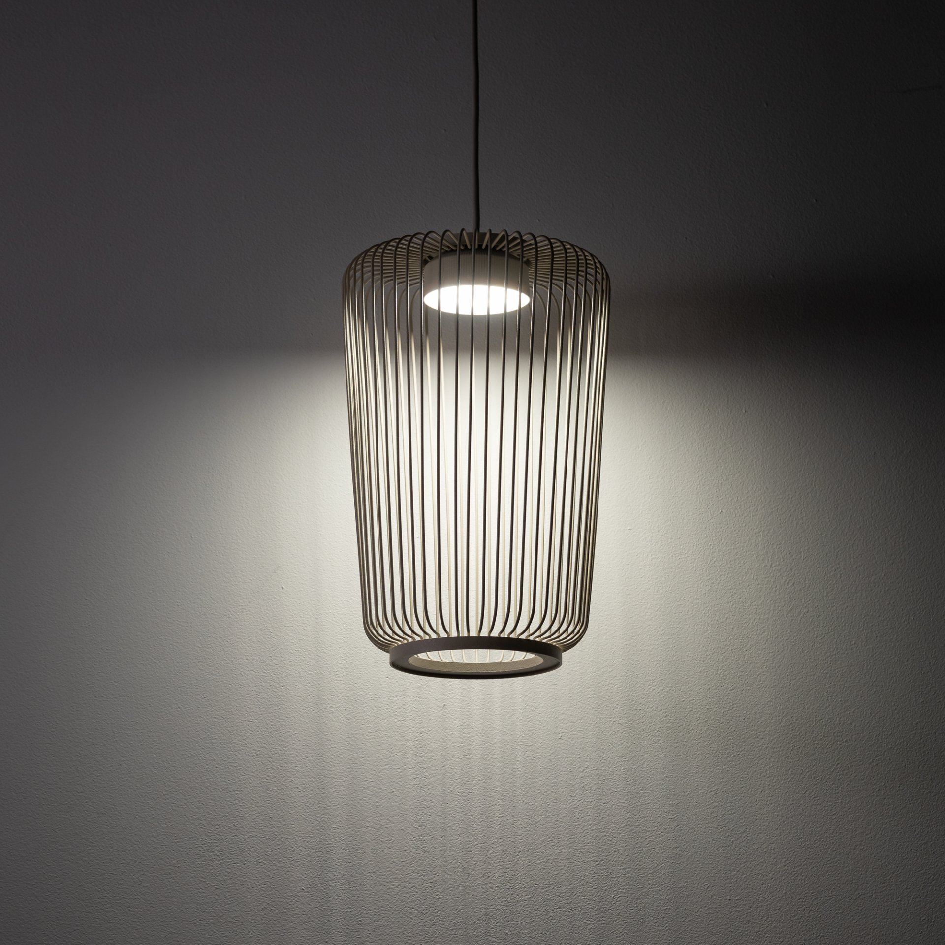 Lampa wisząca z metalowym kloszem w kształcie walca, emitująca światło na ścianę. Klosz składa się z cienkich, równoległych prętów, tworzących ażurową strukturę.