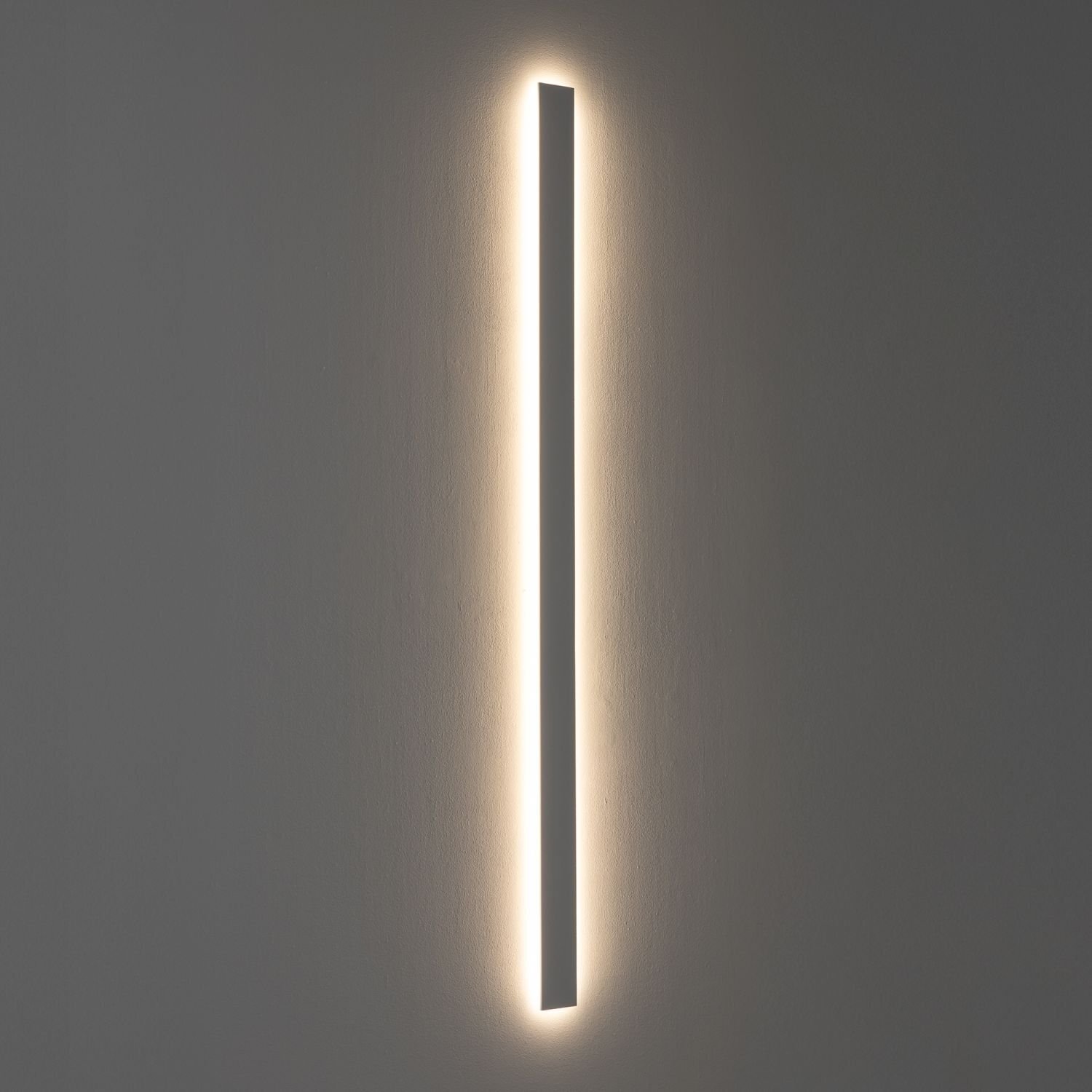 Na obrazie widoczna jest wąska, pionowa lampa ścienna emitująca ciepłe, rozproszone światło na ścianę. Lampa ma prosty, nowoczesny design i jest zamontowana na gładkiej, jednolitej powierzchni.
