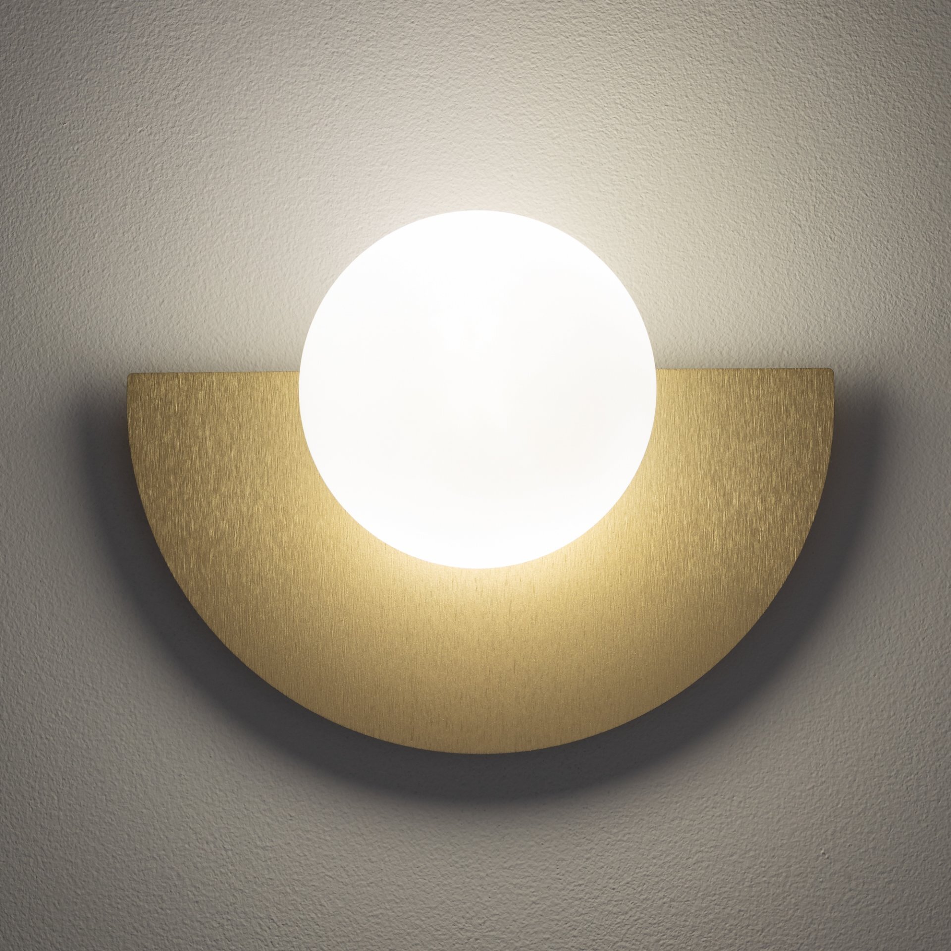 Na obrazie widoczna jest lampa ścienna z okrągłym, białym kloszem umieszczonym na półokrągłej, złotej podstawie. Światło lampy tworzy delikatny efekt świetlny na ścianie.