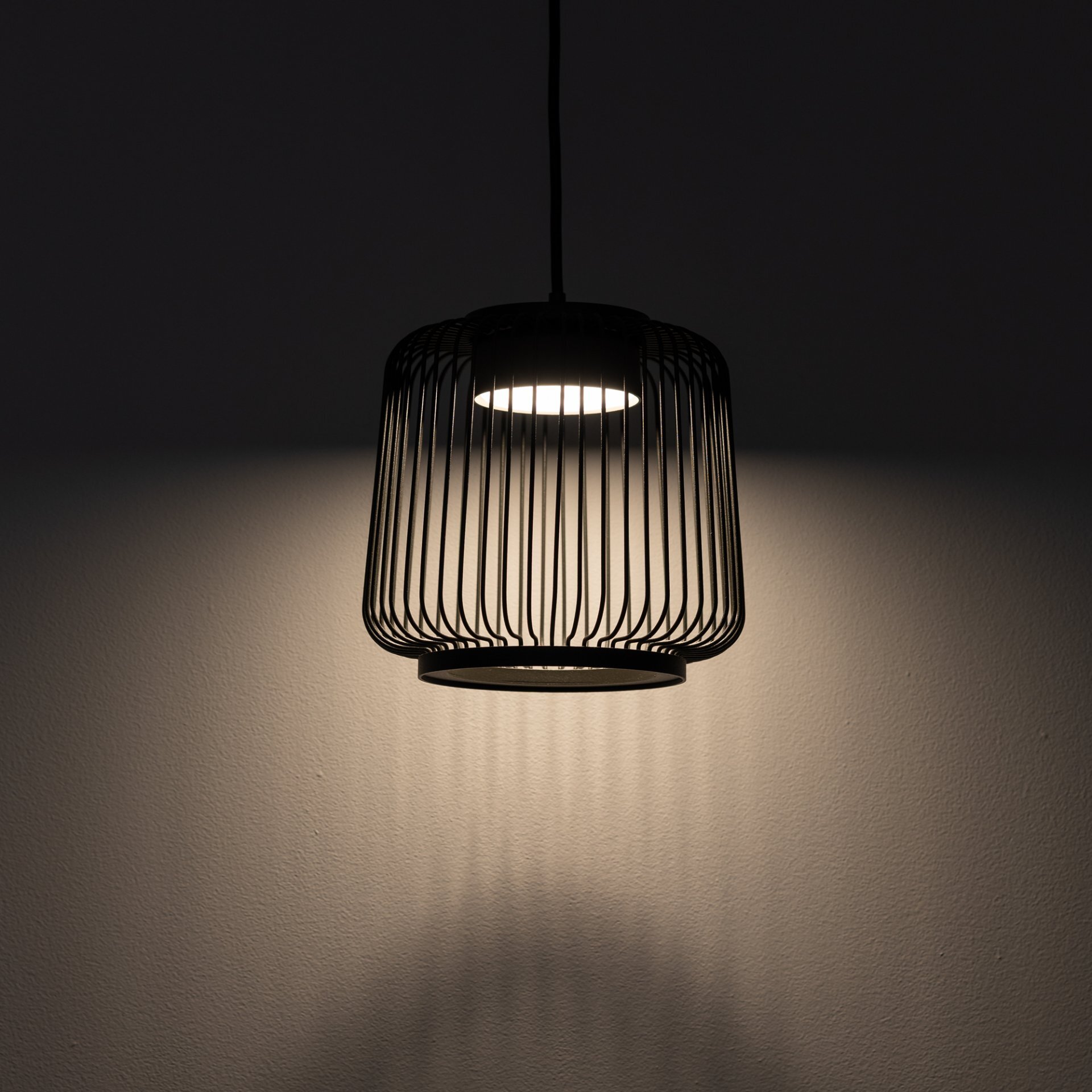 Czarna lampa wisząca z metalowym kloszem w formie ażurowej konstrukcji, emitująca światło na ścianę. Światło tworzy delikatny wzór cieni na powierzchni.