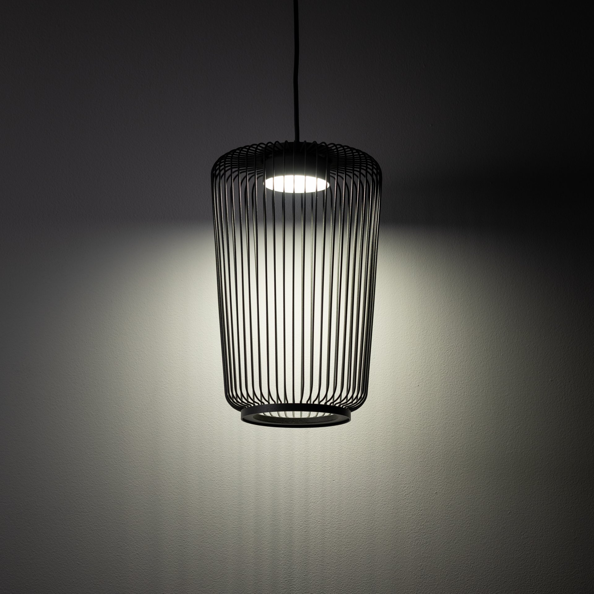 Czarna, wisząca lampa z metalowym kloszem o cylindrycznym kształcie, emitująca światło na ścianę. Światło tworzy efektowne cienie na powierzchni ściany.