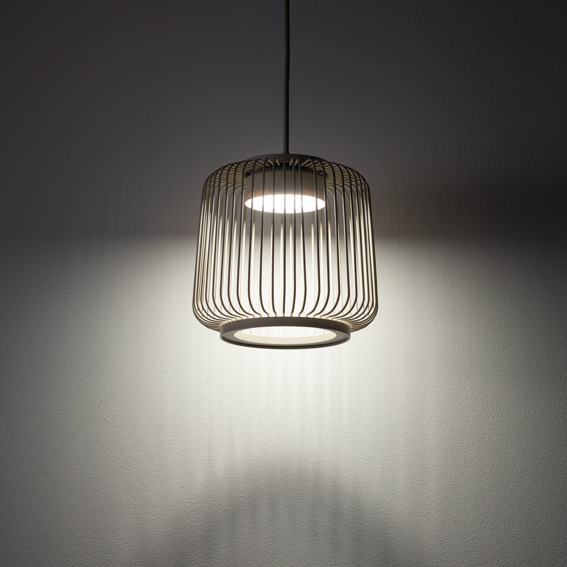 Lampa wisząca z metalowym kloszem w formie ażurowej konstrukcji, emitująca światło w dół na ścianę. Światło tworzy efektowne cienie na powierzchni ściany.