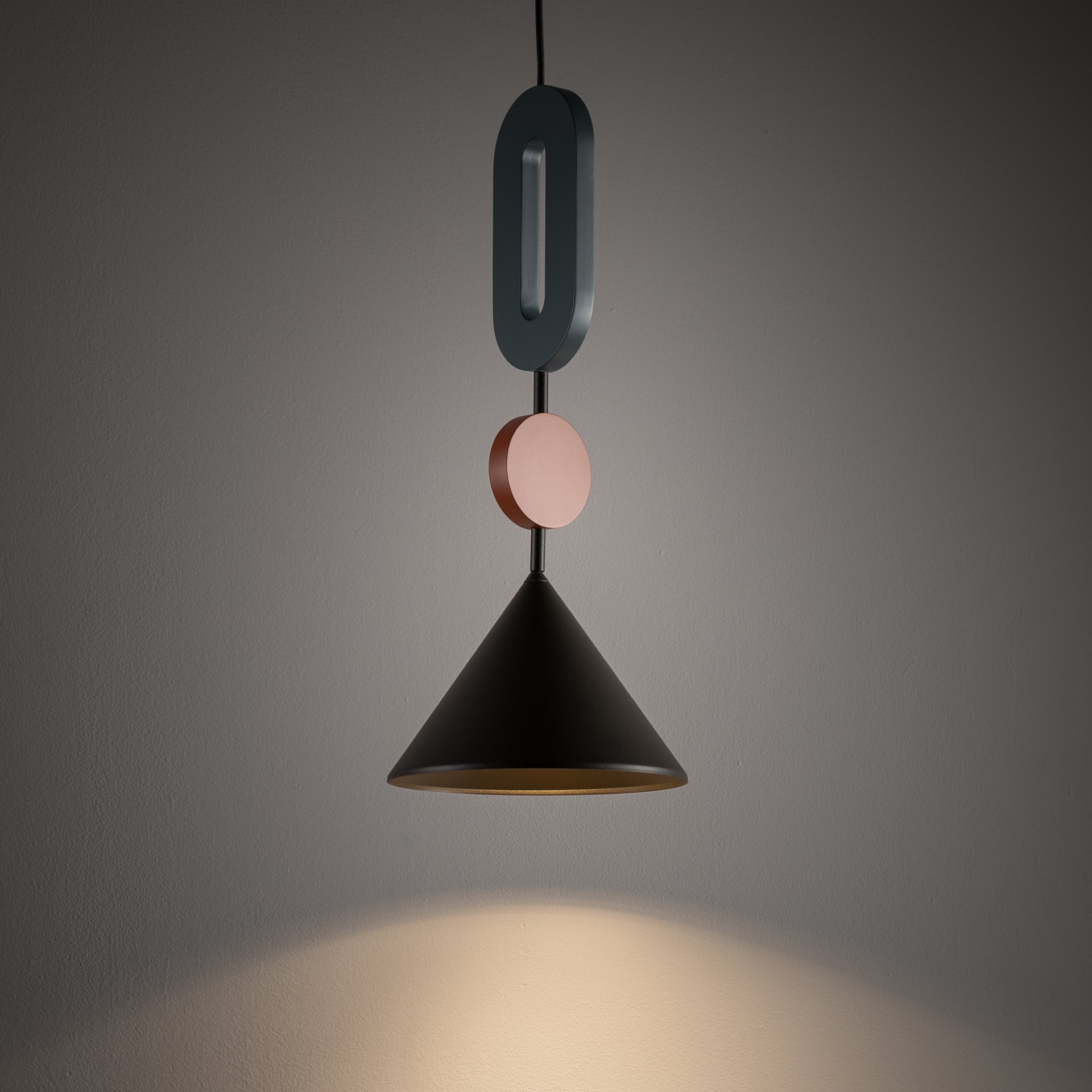 Lampa wisząca o stożkowym kloszu w kolorze czarnym, z ozdobnymi elementami w kształcie owalu i koła w kolorach ciemnoniebieskim i różowym. Światło pada na ścianę, tworząc jasną plamę.