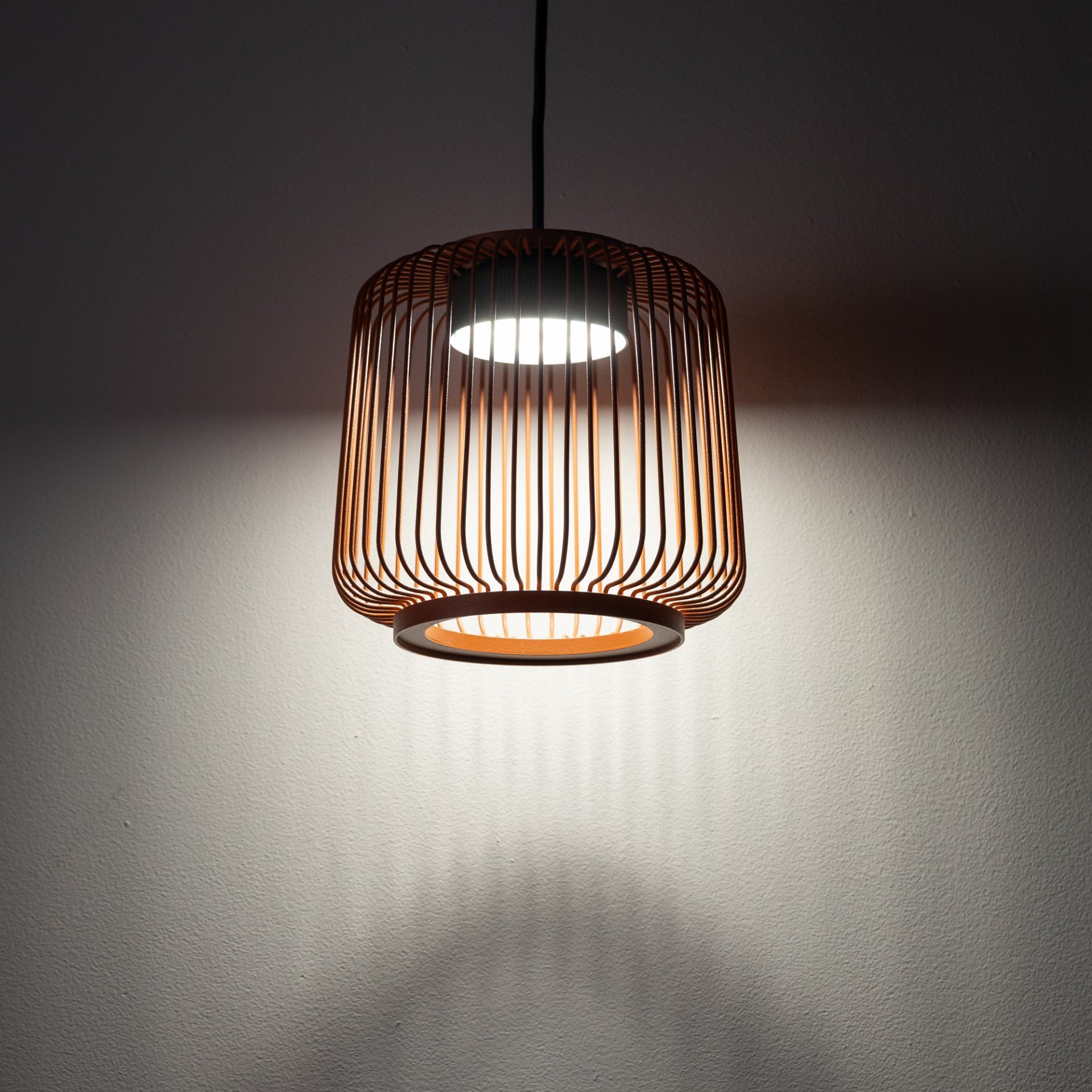 Lampa wisząca z metalowym kloszem w formie ażurowej konstrukcji, emitująca światło na ścianę i podłogę. Klosz ma kolor miedziany, a światło jest białe.
