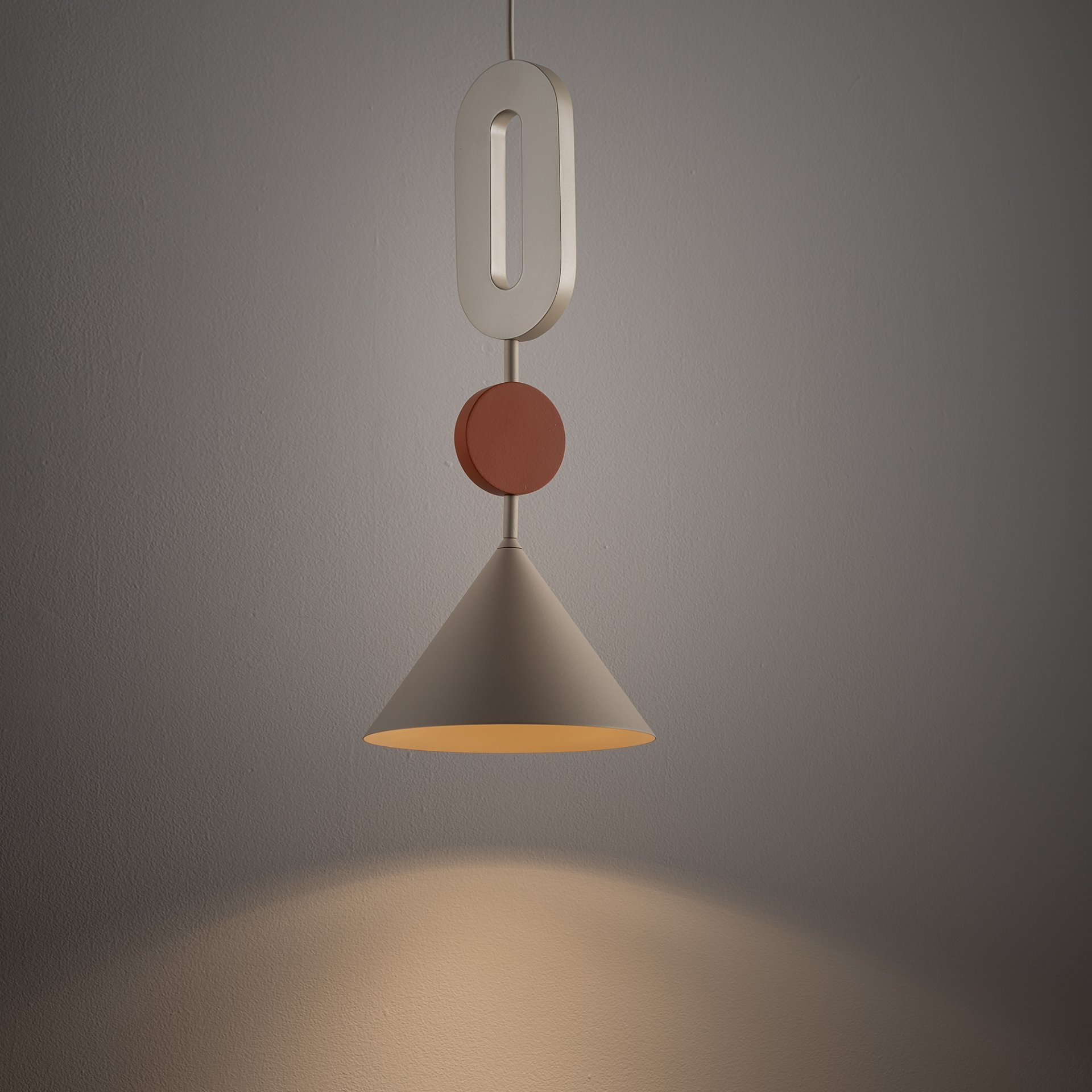 Na obrazie widoczna jest nowoczesna lampa wisząca o stożkowym kloszu, zawieszona na ozdobnym elemencie w kształcie cyfry zero i okrągłym dysku. Lampa emituje ciepłe światło na ścianę, tworząc przytulną atmosferę.