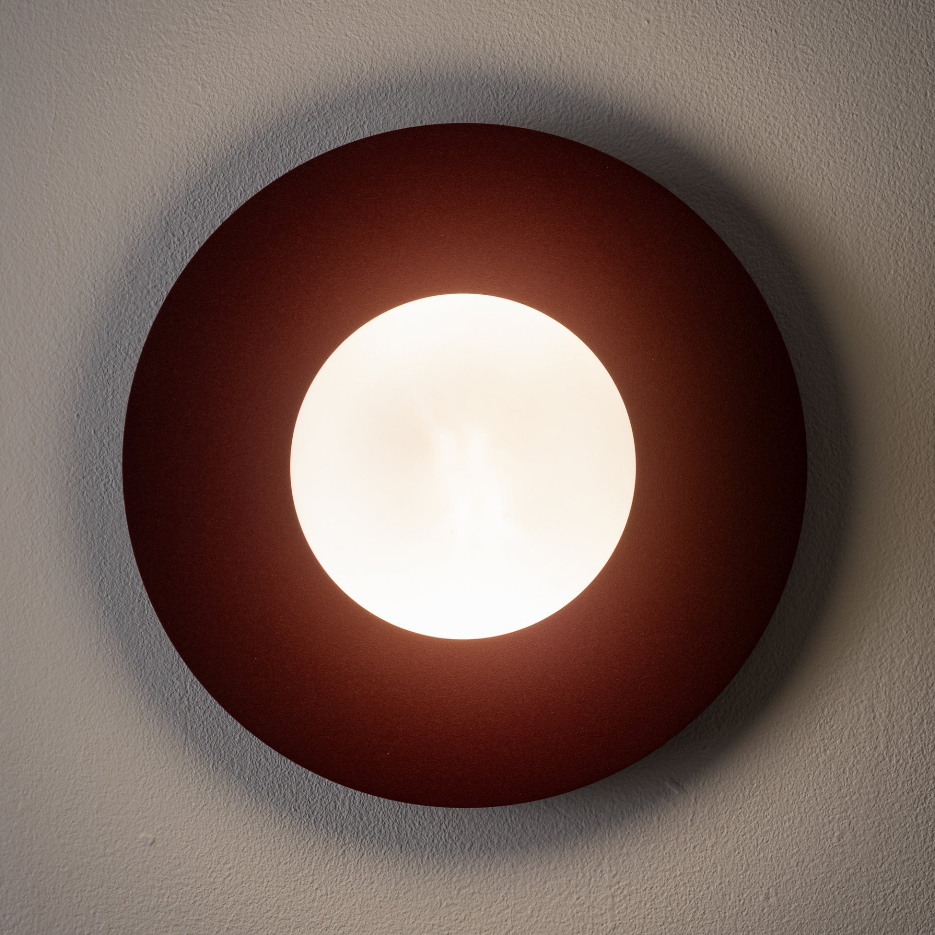 Na obrazie widoczna jest okrągła lampa ścienna z jasnym, centralnym źródłem światła otoczonym ciemniejszym pierścieniem. Lampa jest zamontowana na ścianie.