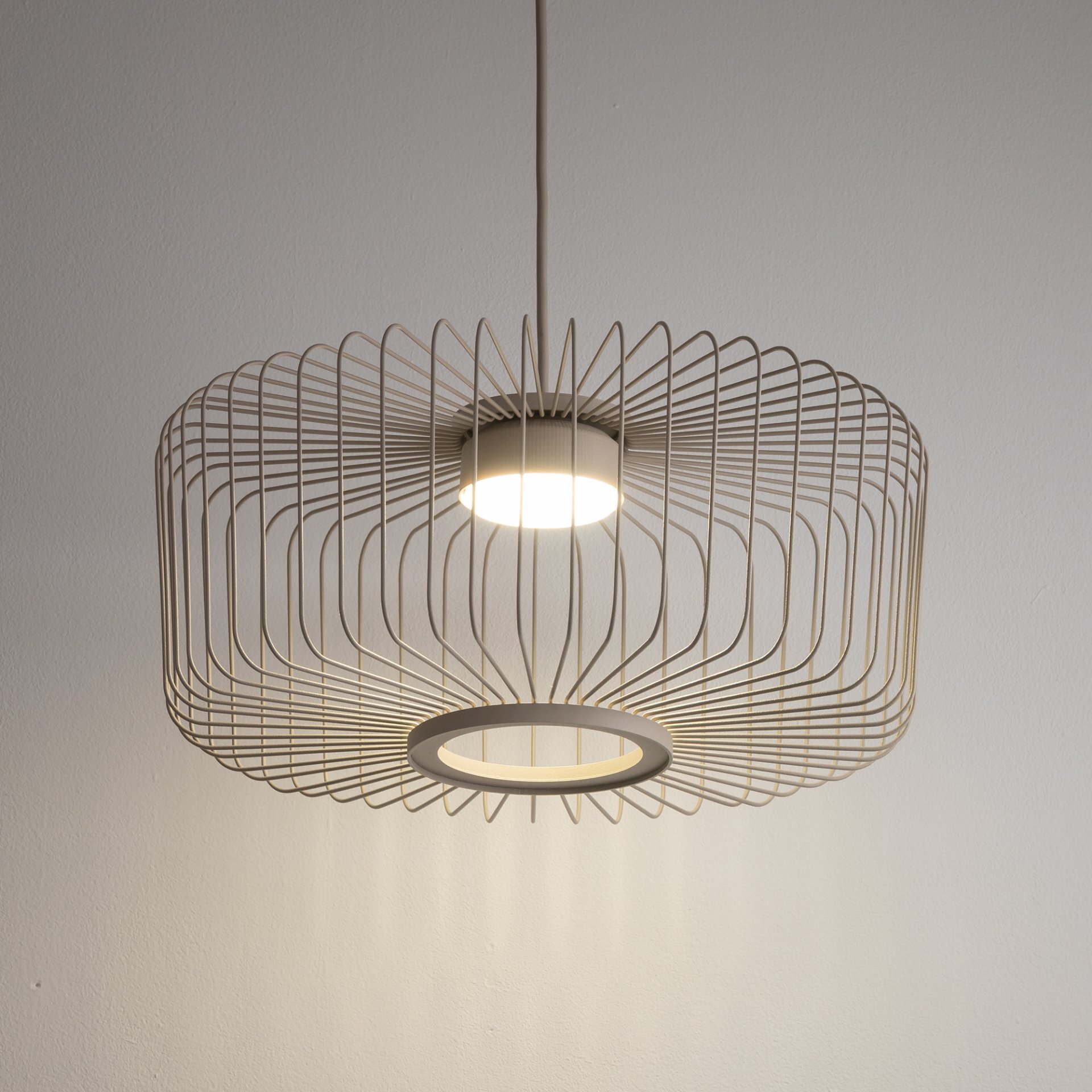 Lampa wisząca z metalowym kloszem o geometrycznym wzorze, emitująca światło z centralnego punktu. Klosz ma jasny kolor i jest zawieszony na cienkim przewodzie.