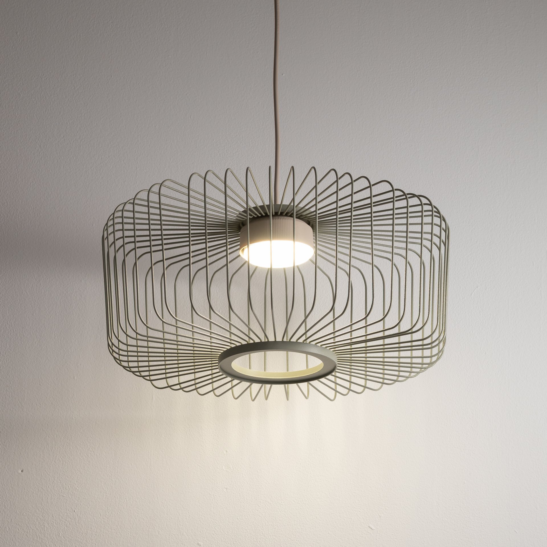 Lampa wisząca z metalowym kloszem o geometrycznym wzorze, zawieszona na przewodzie. Światło emitowane jest z centralnego punktu, tworząc ciekawe cienie na ścianie.