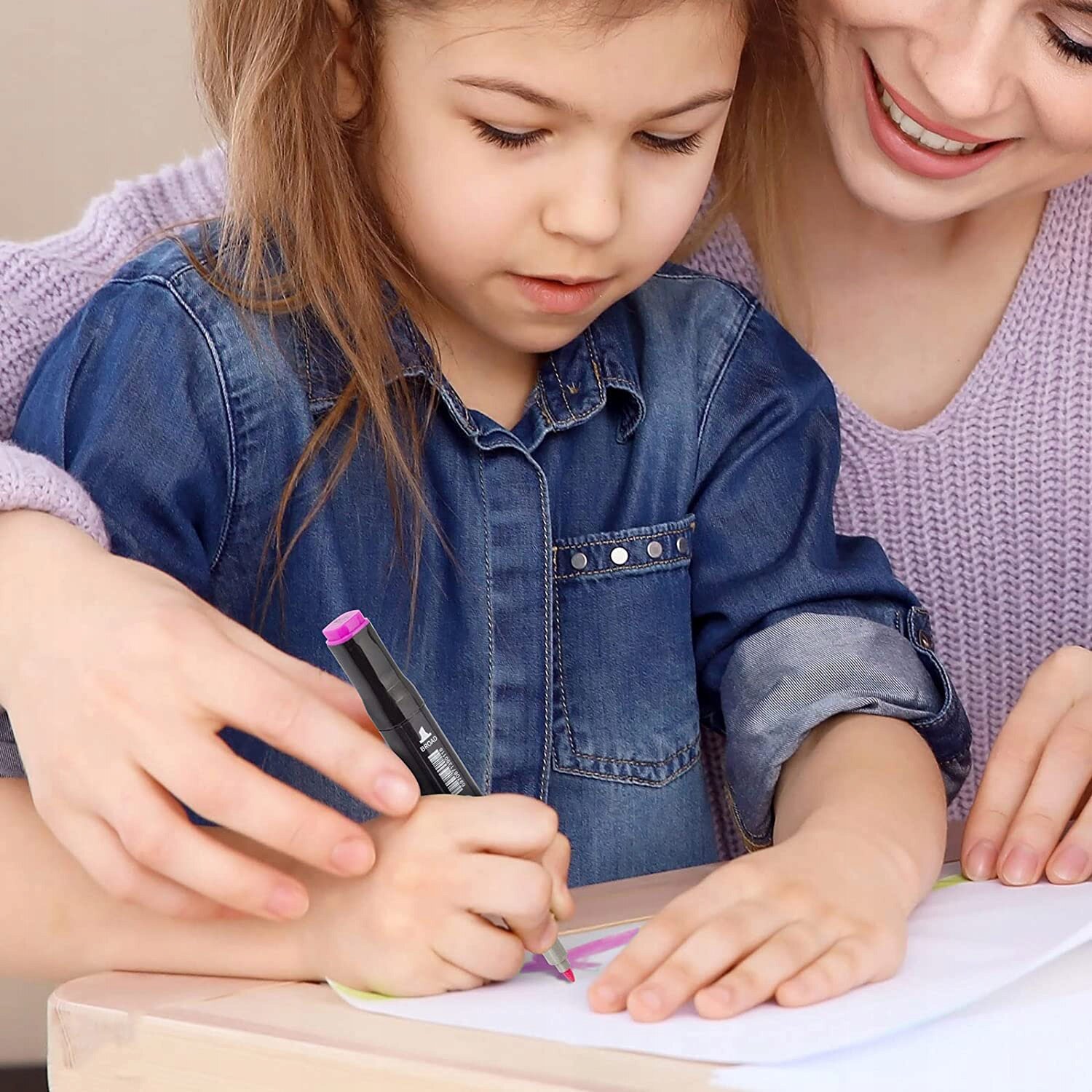 Dziecko rysuje na kartce papieru, trzymając czarny marker z różową końcówką, a obok znajduje się dorosła osoba w fioletowym swetrze.