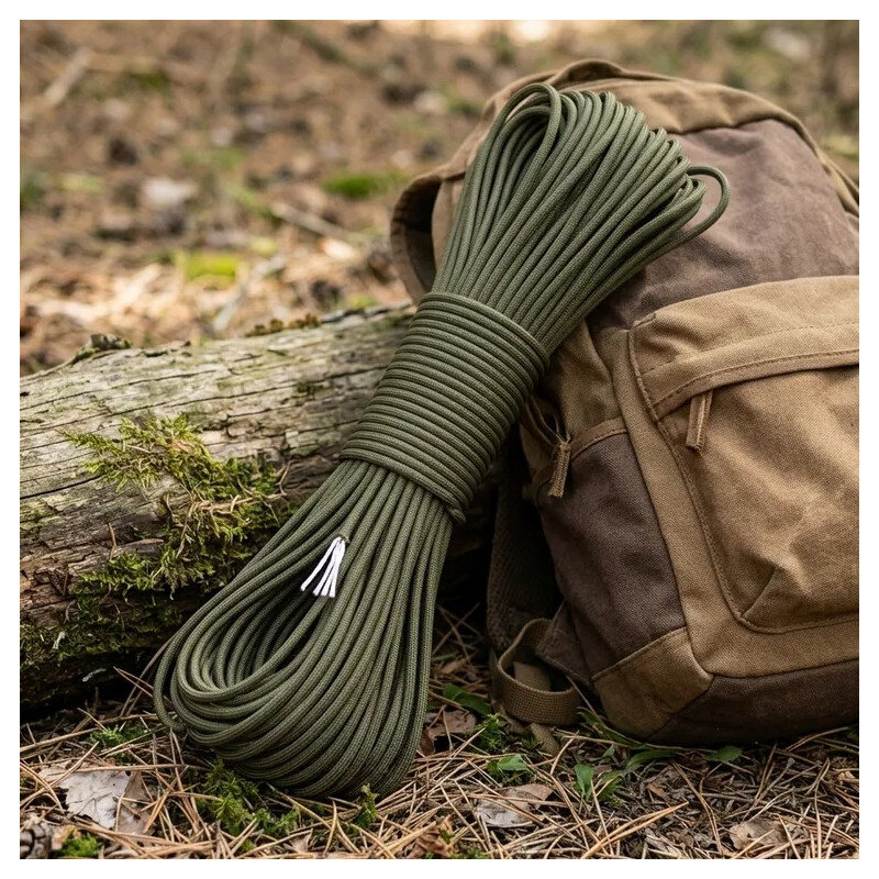 Zielona linka paracord zwinięta w pętlę leży obok brązowego plecaka na tle leśnego podłoża z mchem i liśćmi.