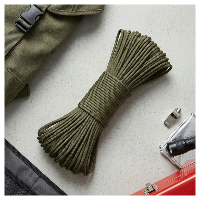 Na obrazie widoczna jest zielona linka paracord zwinięta w pęk, leżąca na jasnej powierzchni. Obok znajdują się elementy takie jak torba, metalowy gwizdek i latarka.