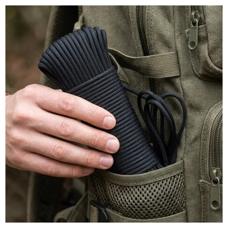 Osoba trzyma czarną linkę paracord, która jest wkładana do kieszeni plecaka z siateczkowym panelem. Plecak ma zamek błyskawiczny i jest wykonany z materiału w kolorze khaki.