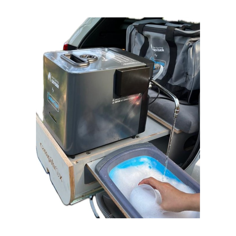 Na zdjęciu znajduje się przenośny podgrzewacz wody z logo \'WATER2GO\', ustawiony na drewnianej platformie. Obok widoczna jest torba oraz ręka trzymająca kubek pod strumieniem wody nad pojemnikiem z pianą.