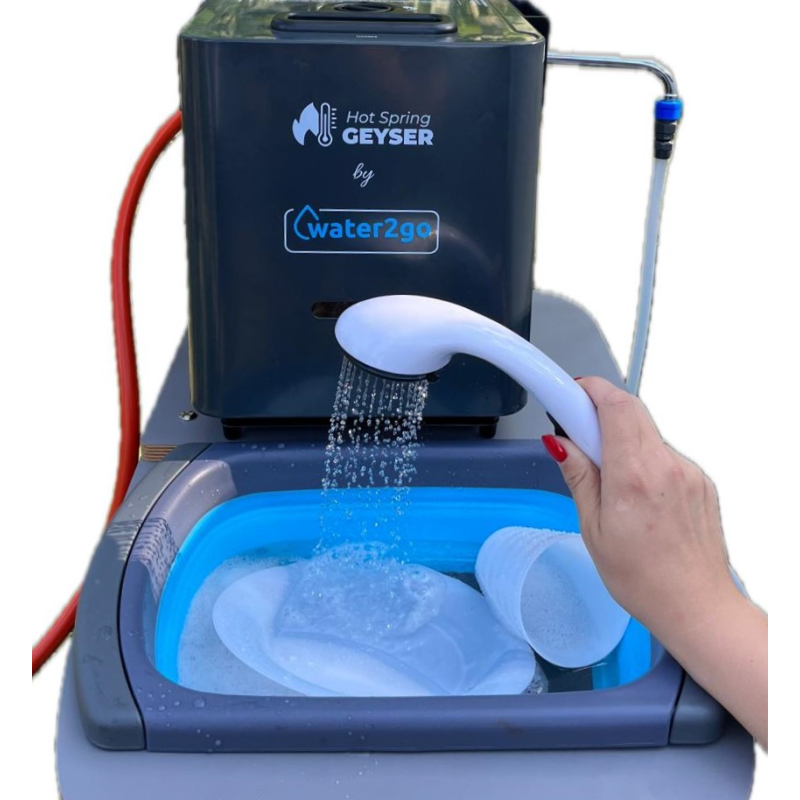 Na obrazie widoczny jest przenośny podgrzewacz wody z napisem \'Hot Spring GEYSER by water2go\'. Osoba trzyma słuchawkę prysznicową, z której leci woda do niebieskiej miski z naczyniami.