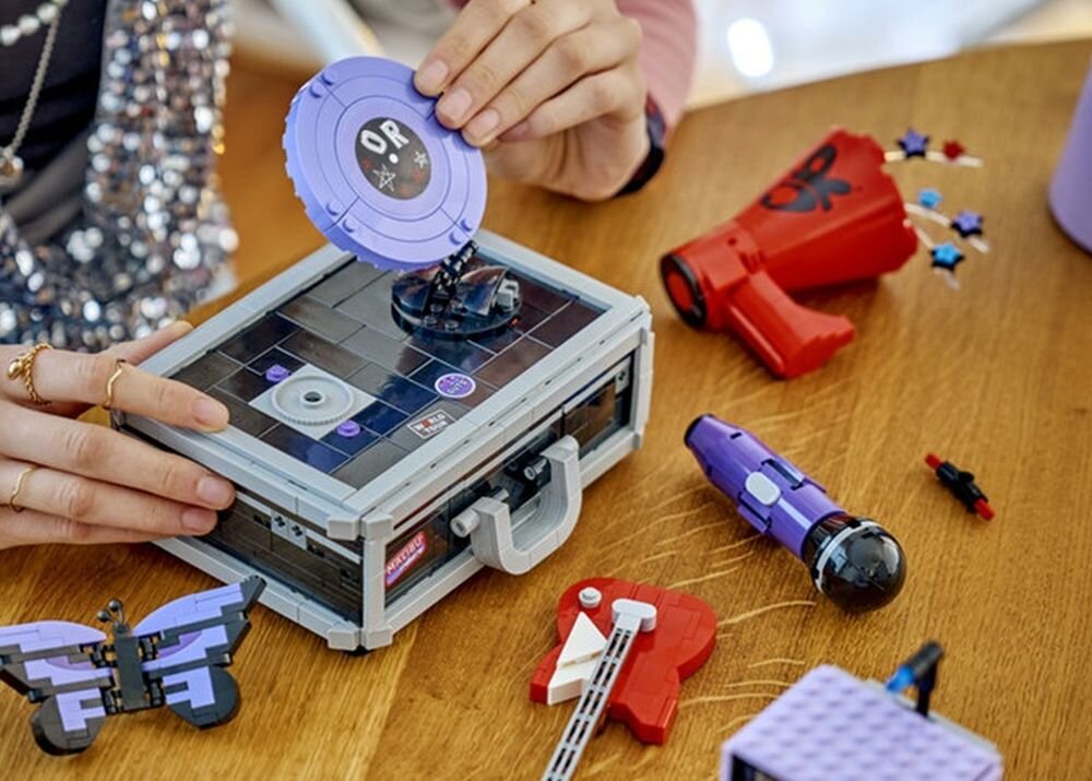 Na drewnianym stole znajduje się zestaw klocków LEGO, w tym pudełko z otwieranym wieczkiem, czerwony megafon, fioletowy motyl i inne akcesoria. Na wieczku pudełka widoczny jest napis \'OR\'.