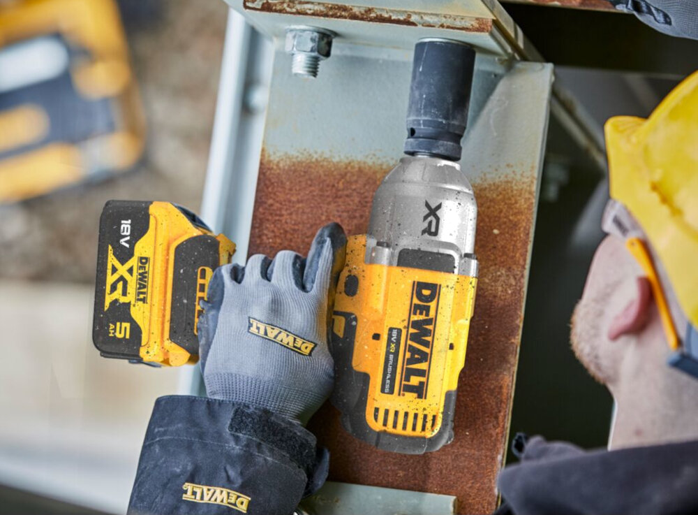 Osoba w kasku używa żółtego klucza udarowego z widocznym logo DEWALT do pracy na metalowej konstrukcji. Obok znajduje się akumulator z oznaczeniem 18V XR 5 Ah.