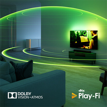 W salonie znajduje się telewizor wyświetlający obraz z zielonymi efektami świetlnymi wokół niego. Na dole obrazu widoczne są loga \'Dolby Vision Atmos\' i \'dts Play-Fi\'.