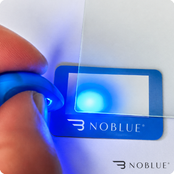 Osoba trzyma przezroczysty filtr, przez który przechodzi niebieskie światło. Na obrazie widoczne jest logo \'NOBLUE\'.
