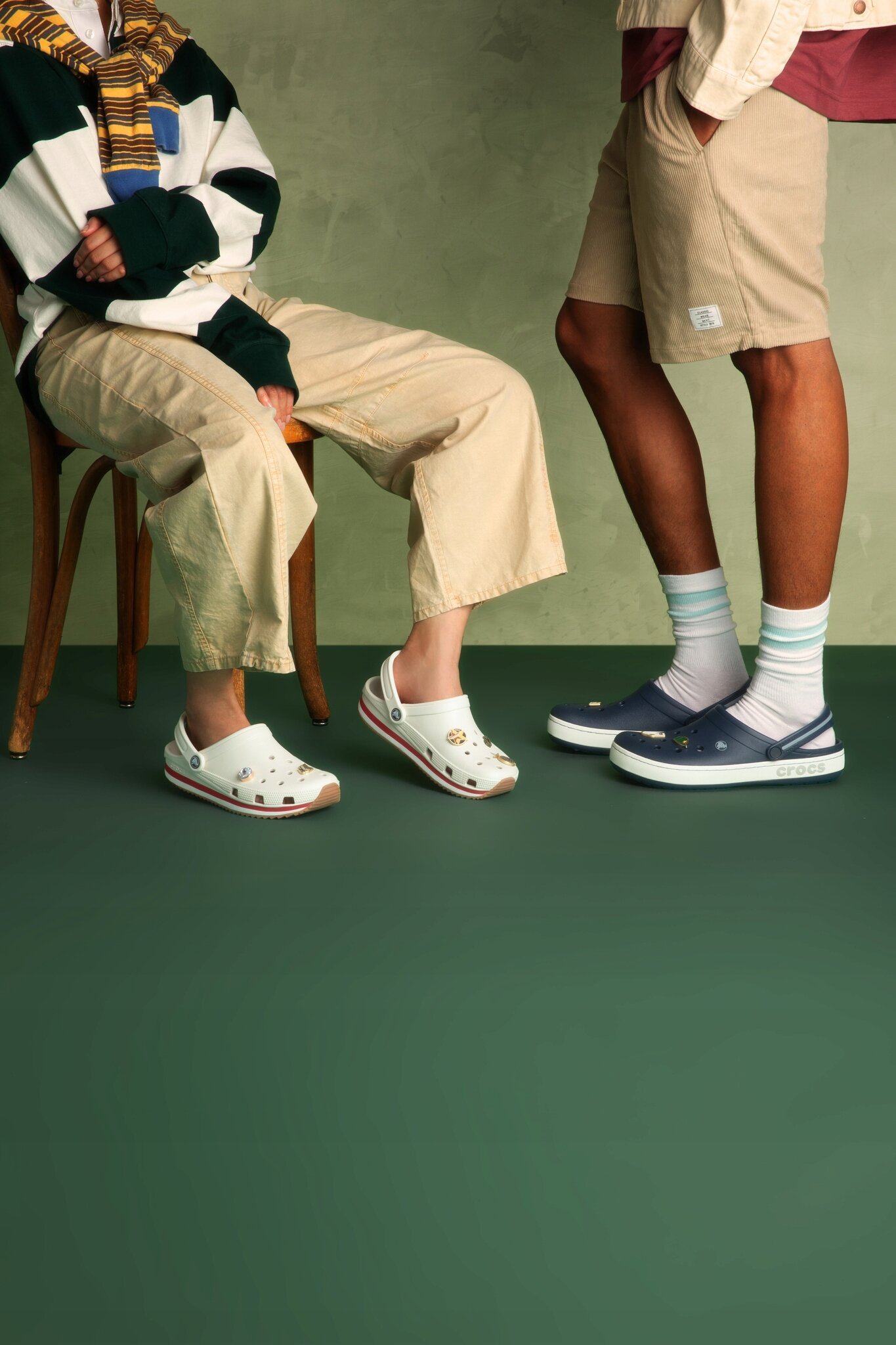 Na zdjęciu widać dwie osoby siedzące na krzesłach, noszące różne modele butów Crocs. Jedna osoba ma białe chodaki z ozdobami, a druga granatowe z białą podeszwą.