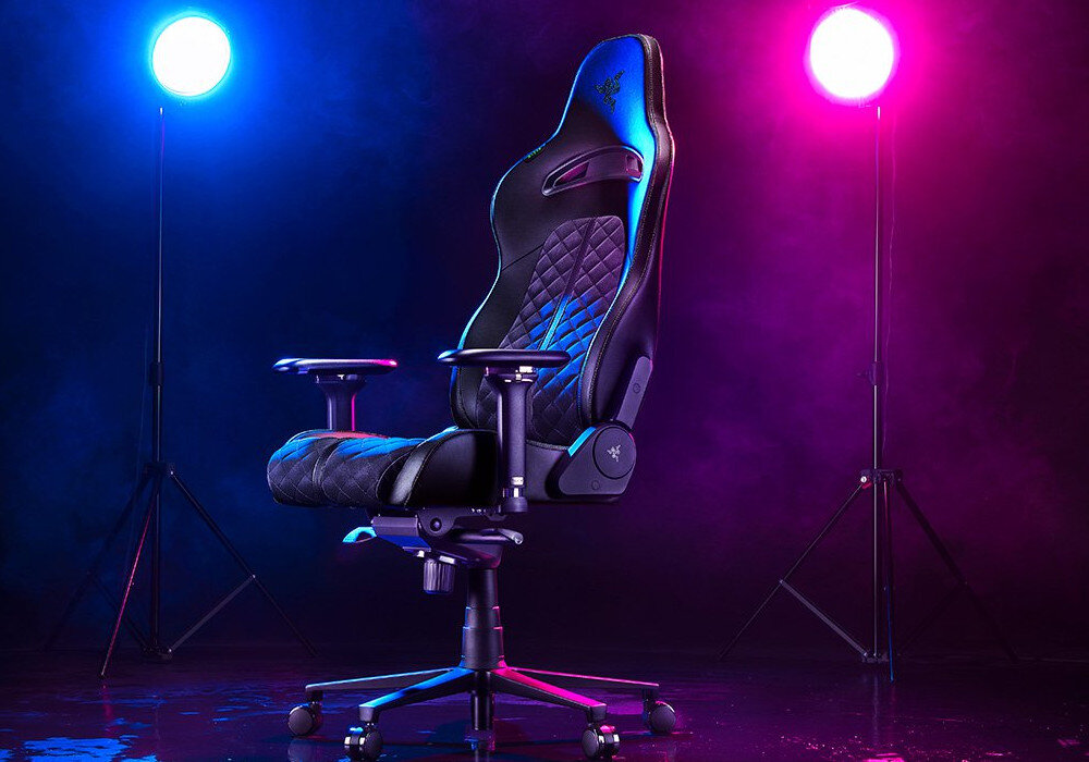 Czarny fotel gamingowy z logo Razer na oparciu, ustawiony na tle kolorowych świateł w odcieniach niebieskiego i fioletowego. Fotel ma pikowane siedzisko i regulowane podłokietniki.