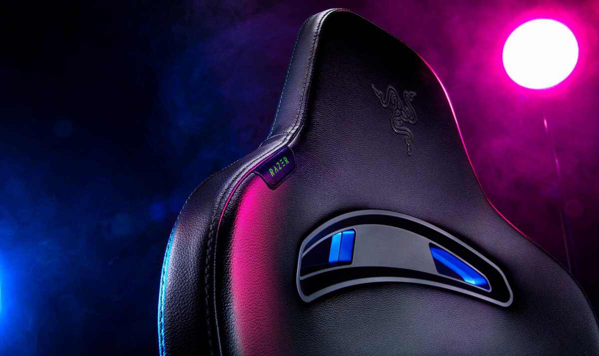 Czarny fotel gamingowy z logo RAZER na oparciu, oświetlony kolorowymi światłami w tle.