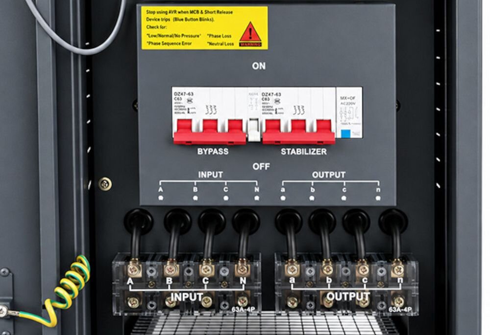 Panel sterowania stabilizatora napięcia z widocznymi przełącznikami oznaczonymi jako BYPASS, STABILIZER oraz sekcjami INPUT i OUTPUT. Na górze znajduje się żółta naklejka z ostrzeżeniem i instrukcjami dotyczącymi urządzenia.
