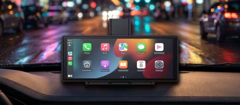 Na desce rozdzielczej samochodu znajduje się tablet z wyświetlaczem, na którym widoczne są ikony aplikacji takich jak Phone, Music, Maps, Messages, Now Playing, SmartDevice, Podcasts, Audiobooks, Calendar i Settings. W tle widać rozmyte światła uliczne, sugerujące jazdę nocą.