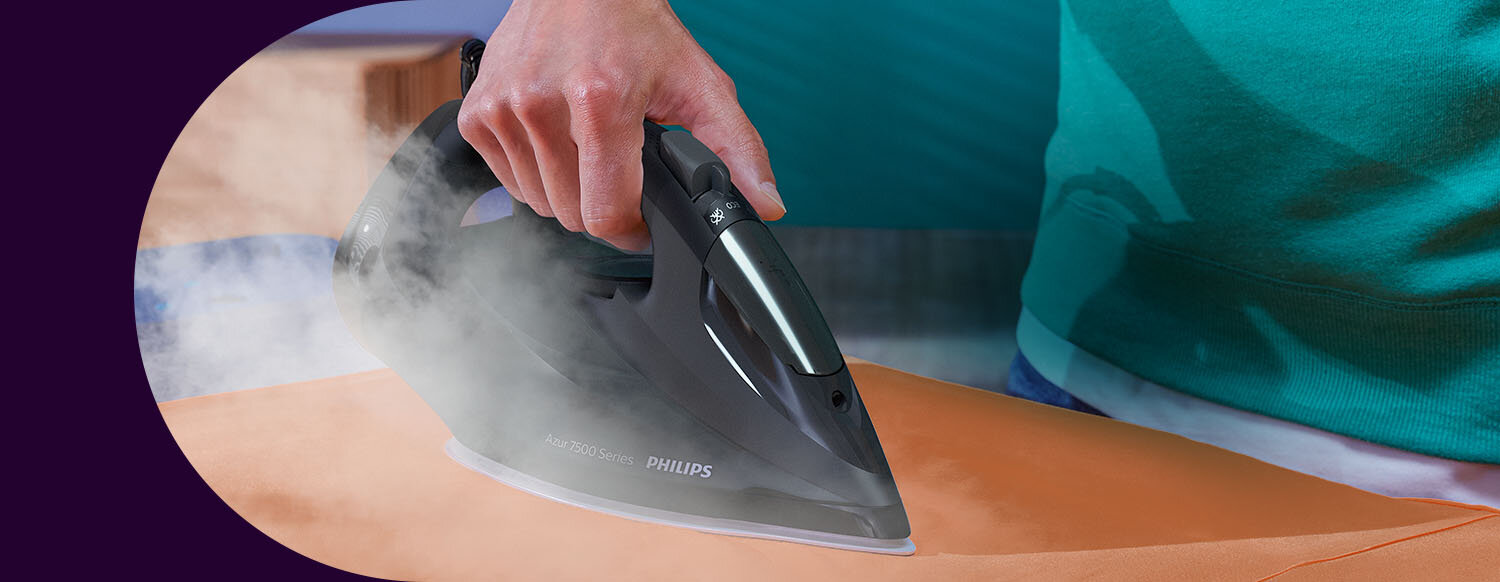 Osoba prasuje pomarańczowy materiał żelazkiem marki PHILIPS, z którego wydobywa się para. Na żelazku widoczny jest napis \'PHILIPS\'.