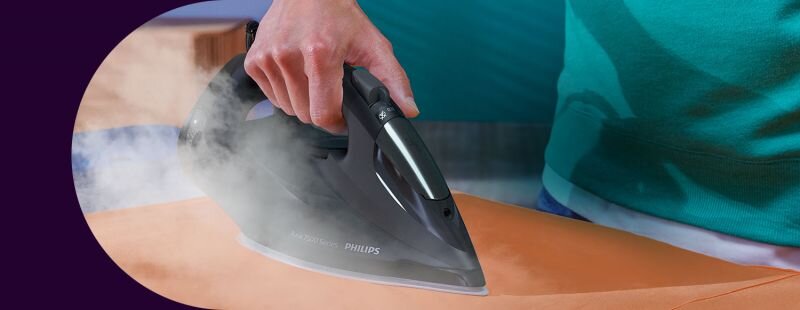 Osoba prasuje pomarańczowy materiał żelazkiem marki PHILIPS, z którego wydobywa się para. Na żelazku widoczny jest napis \'PHILIPS\'.