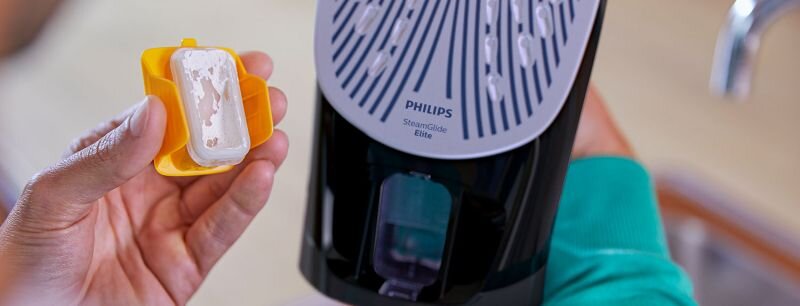 Osoba trzyma żółty pojemnik z białym osadem obok żelazka z widoczną stopą oznaczoną napisem \'PHILIPS SteamGlide Elite\'.