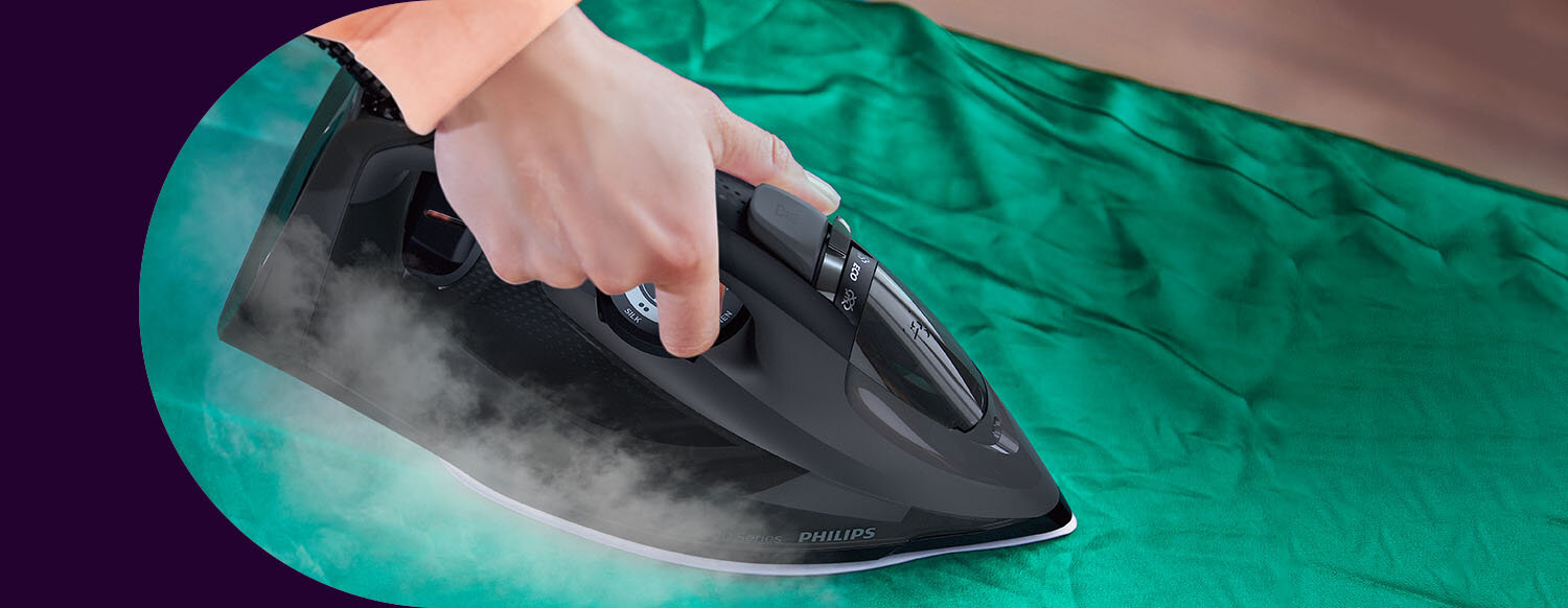 Osoba prasuje zielony materiał czarnym żelazkiem z widocznym logo \'PHILIPS\'. Z żelazka wydobywa się para.