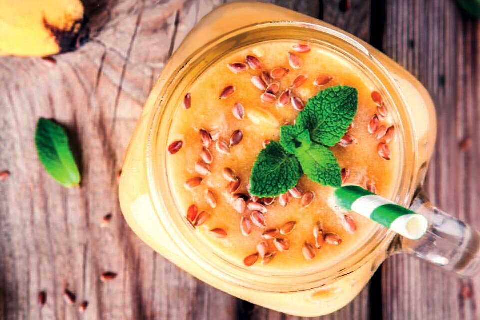 Szklanka z pomarańczowym smoothie, udekorowana nasionami i listkiem mięty, z zielono-białą słomką. Tło stanowi drewniana powierzchnia.