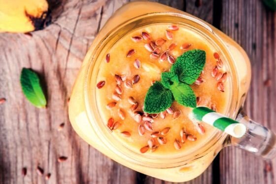 Szklanka wypełniona pomarańczowym smoothie, ozdobiona listkami mięty i posypana nasionami, z zielono-białą słomką. Tło stanowi drewniana powierzchnia.