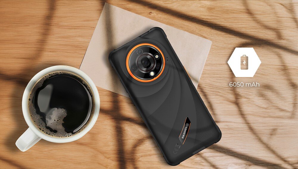 Na drewnianym stole leży smartfon z widocznym logo \'HAMMER\', obok filiżanki kawy. Obok telefonu znajduje się ikona baterii z napisem \'6050 mAh\'.