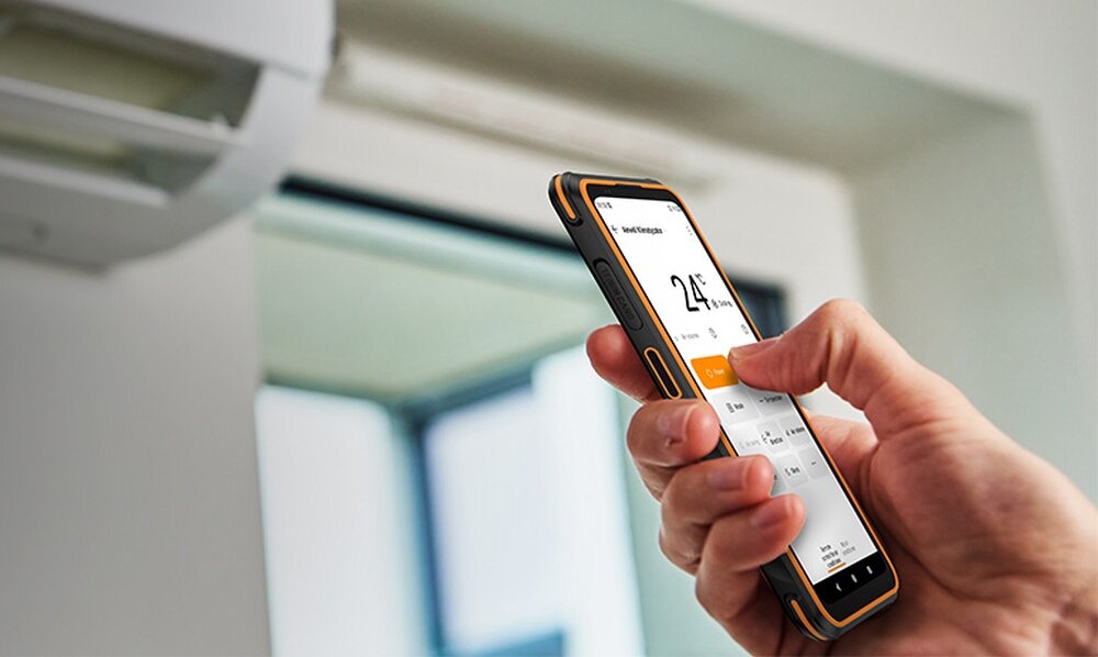 Osoba trzyma pomarańczowy smartfon, na ekranie widoczna jest aplikacja z ustawieniami temperatury 24°C. W tle znajduje się klimatyzator.