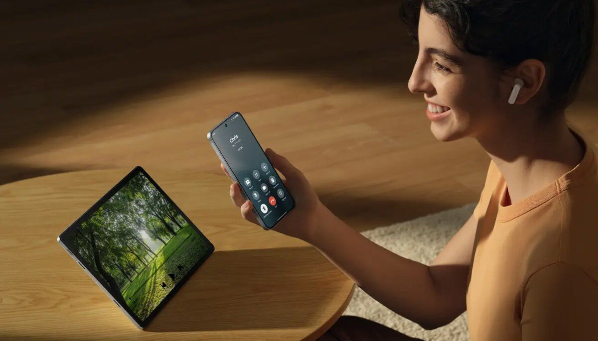 Osoba trzyma smartfon z widocznym ekranem połączenia telefonicznego, a na stoliku obok znajduje się tablet z wyświetlonym obrazem lasu. Osoba ma w uchu białe słuchawki douszne.