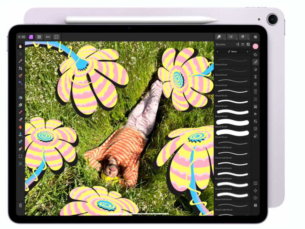 Tablet z wyświetlaczem pokazującym aplikację graficzną z kolorowymi kwiatami i osobą leżącą na trawie. Na ekranie widoczne są narzędzia do rysowania oraz różne pędzle.