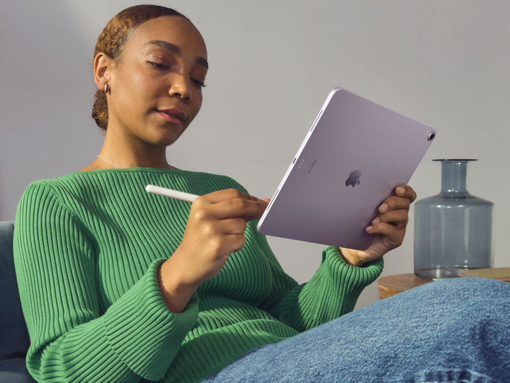 Osoba trzyma tablet w kolorze niebieskim z logo APPLE, używając rysika. W tle widoczny jest szklany wazon na drewnianym stoliku.