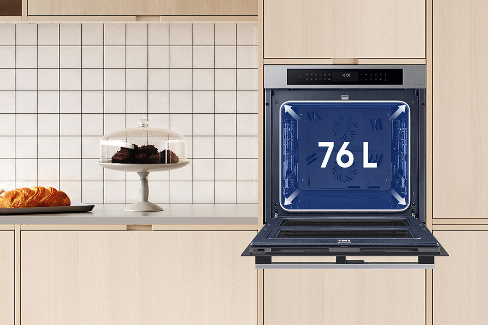 Piekarnik Samsung NV7B4345VAS w zabudowie meblowej z otwartymi drzwiami pokazuje jak pojemna jest komora Otwarty piekarnik w zabudowie kuchennej z widoczną pojemnością 76 L na drzwiach. Obok na blacie znajduje się patera z ciastem i talerz z pieczywem.