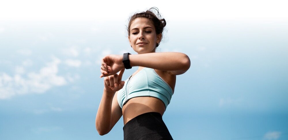 Osoba w sportowym stroju patrzy na smartwatch na tle nieba. Smartwatch ma ciemny pasek.